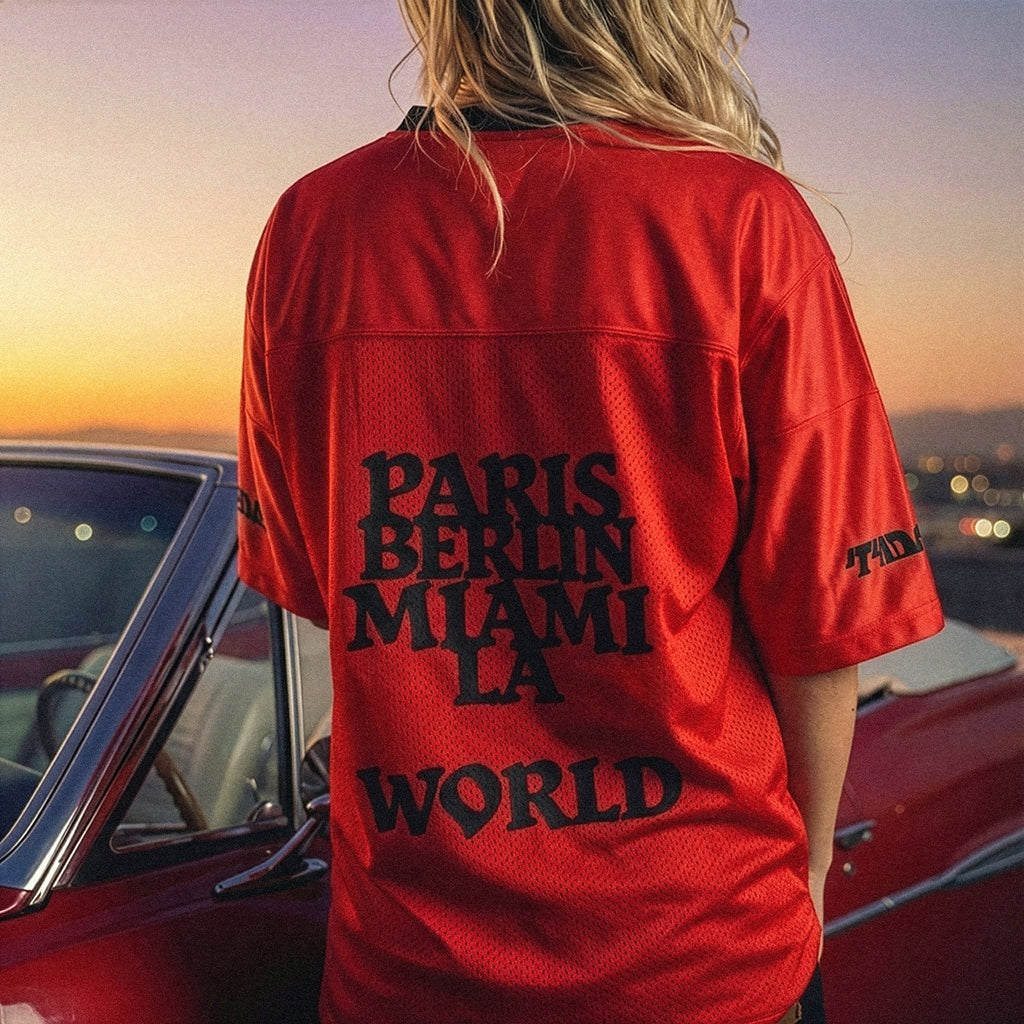 Person wearing a red jersey with city names printed on it, standing in front of a classic car at sunset.